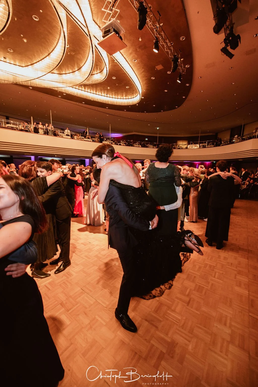 Tanzende Paare auf einem festlichen Ball in einem großen Saal mit gläserner Decke und mehreren Ebenen.