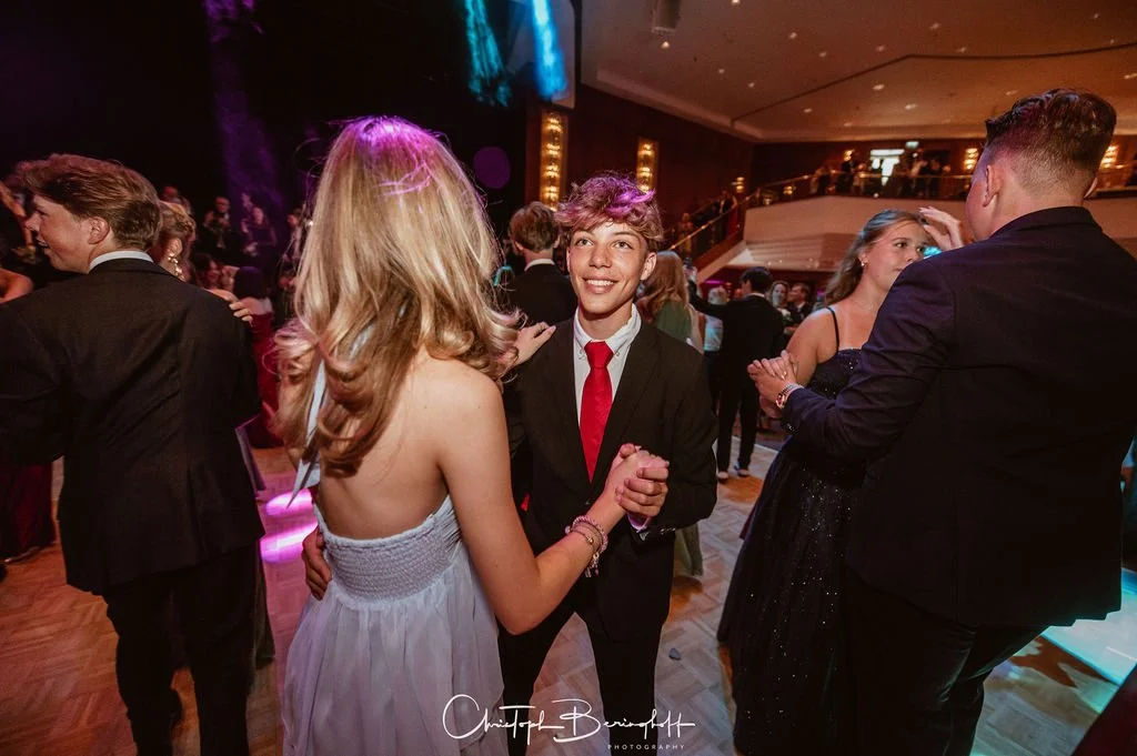 Jugendliche tanzen auf einer Party im festlichen Abendkleid und Anzug in einem eleganten Saal.