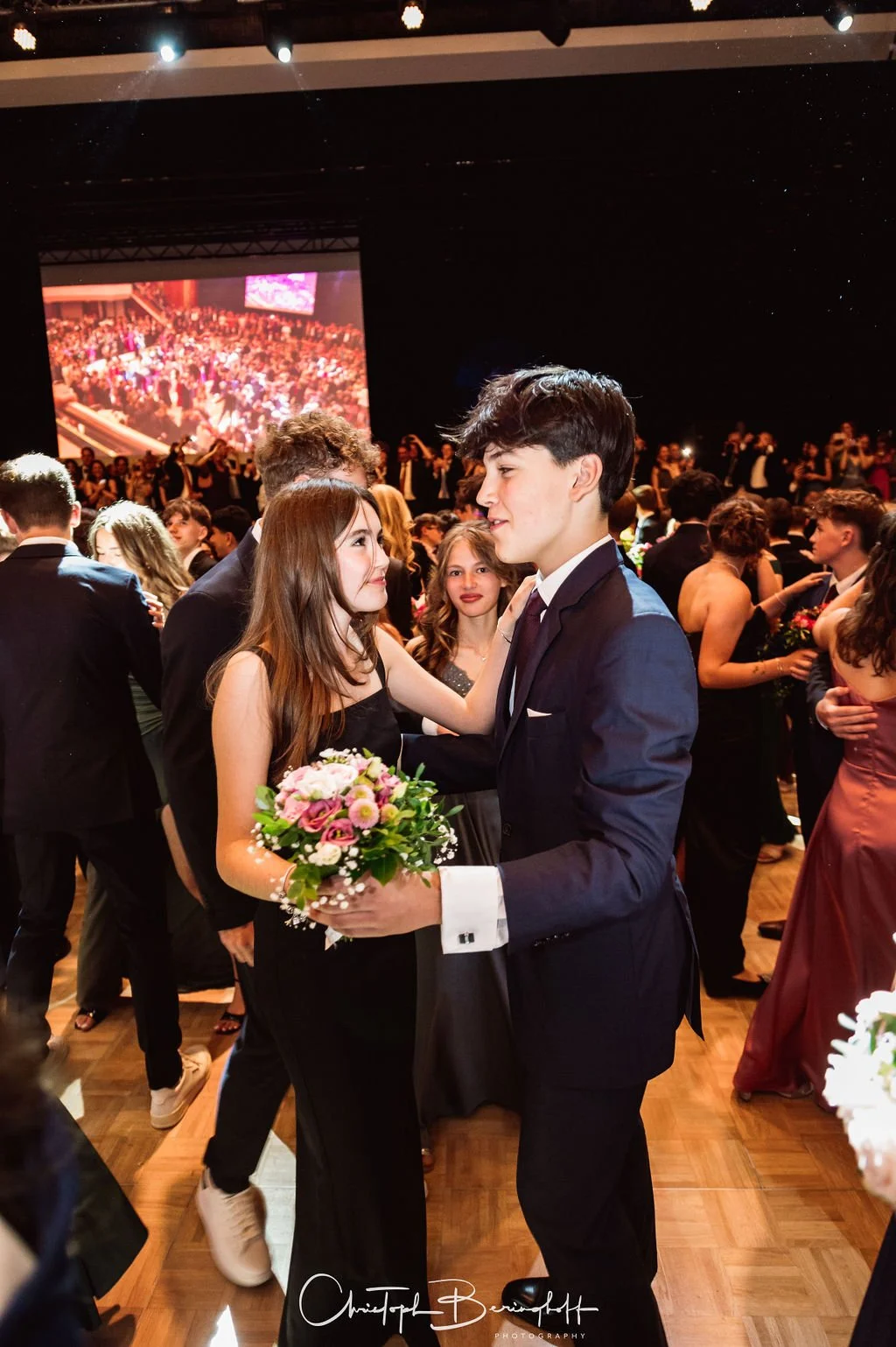 Junge Männer und Frauen bei einer formellen Tanzveranstaltung, welche in einem großen Saal mit Bühne und Leinwand im Hintergrund stattfinden.