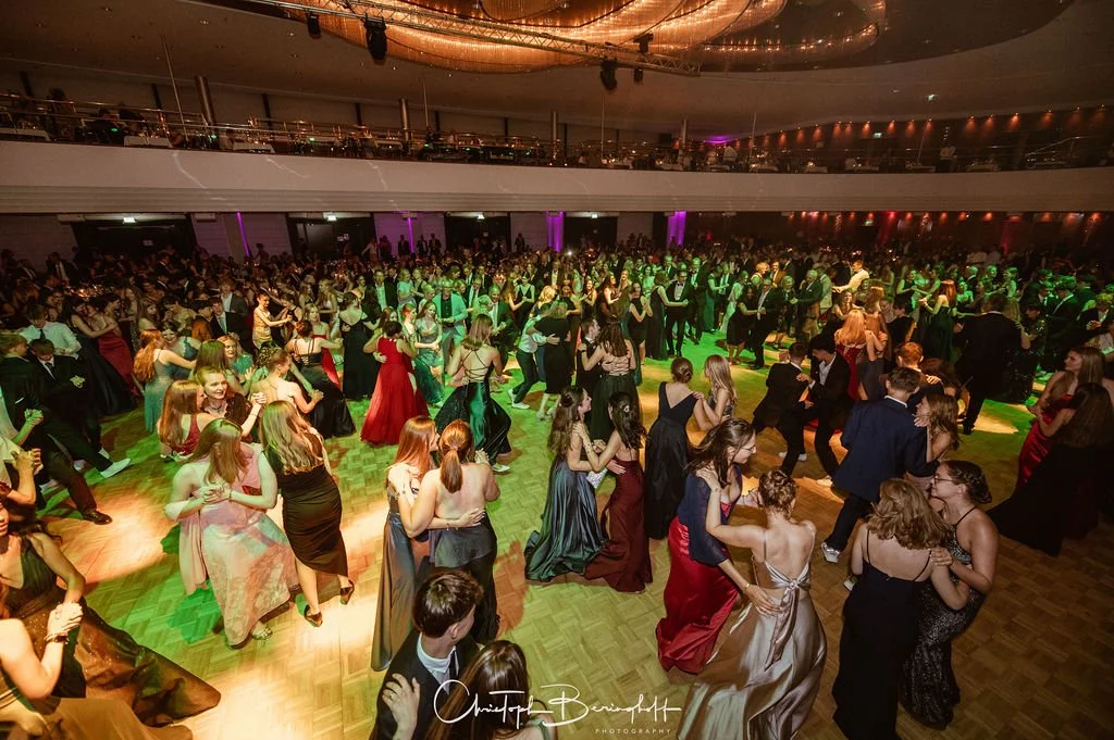 Menschen tanzen auf einer festlich beleuchteten Ballsaal-Tanzfläche, umgeben von mehreren Gästen in eleganten Abendkleidern und Anzügen.