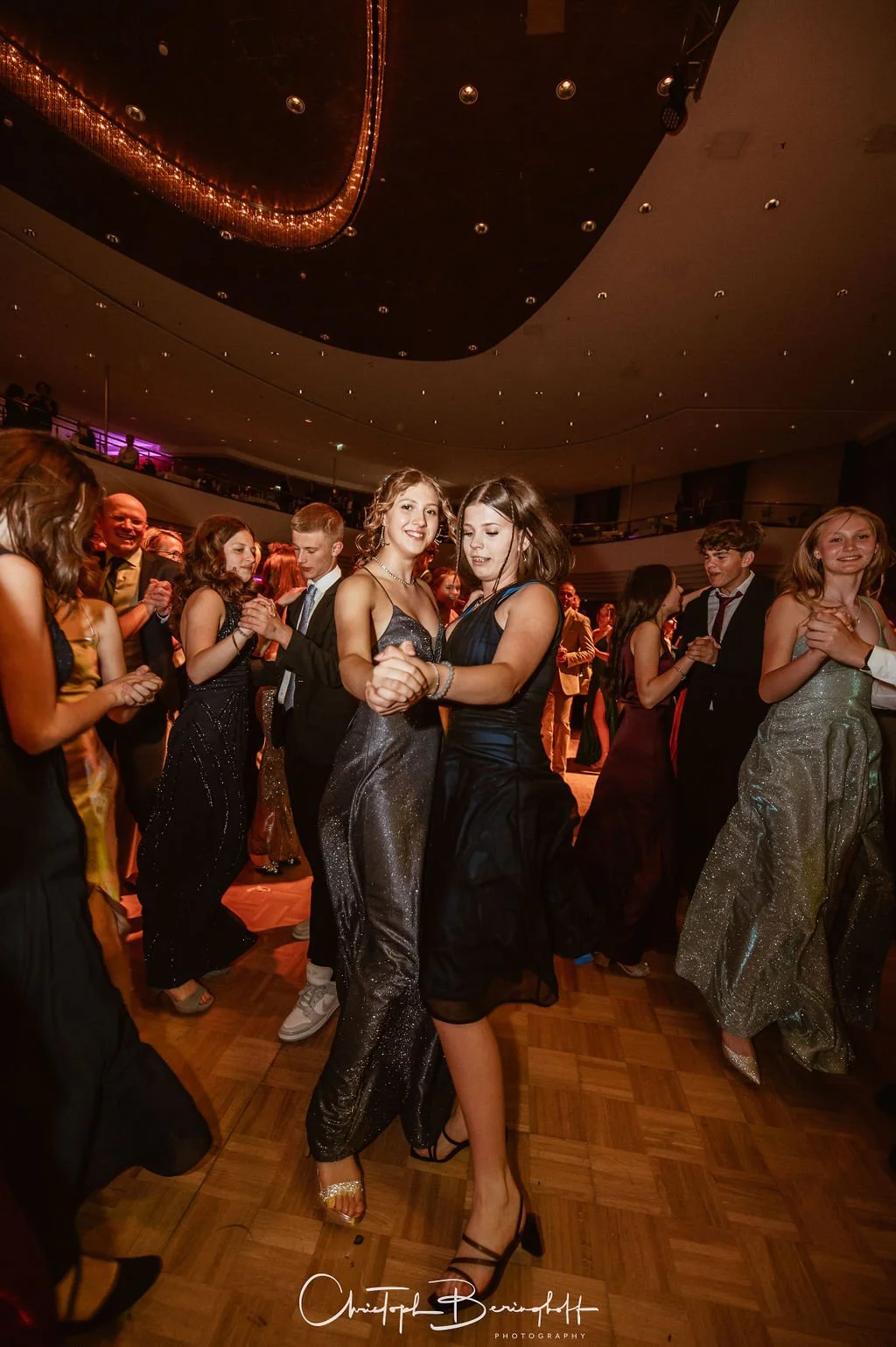 Junge Leute tanzen auf einer Party, alle tragen elegante Abendkleidung, in einem großen Veranstaltungsraum mit Holzparkett und dunkler Decke.