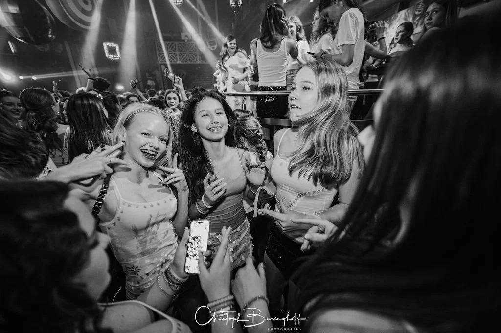 A group of young women in a nightclub, surrounded by other partygoers, with a DJ setup and lighting effects in the background.