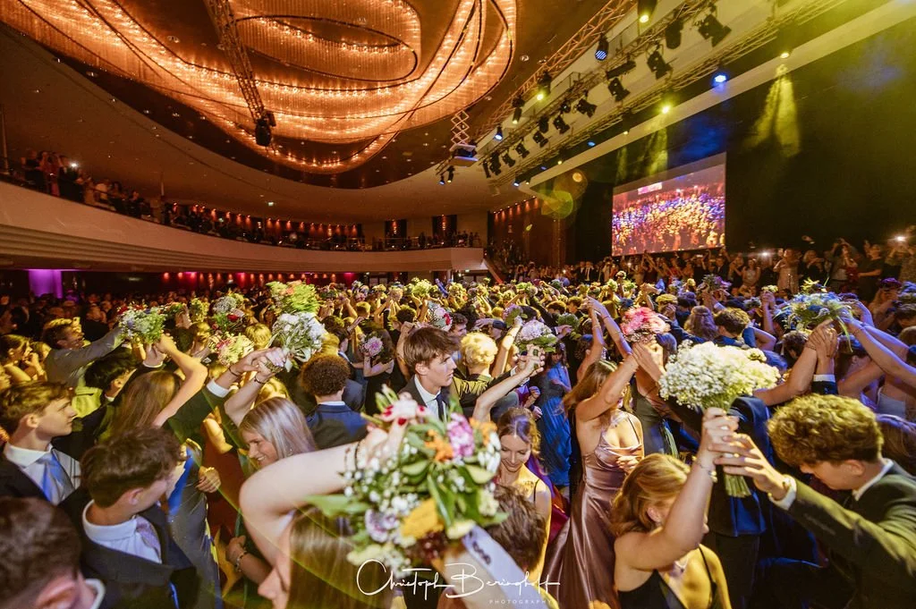 Menschen bei einer Feier im Festsaal, viele tragen Blumenkränze und feiern gemeinsam, mit einer großen Leinwand und bunten Lichteffekten im Hintergrund.