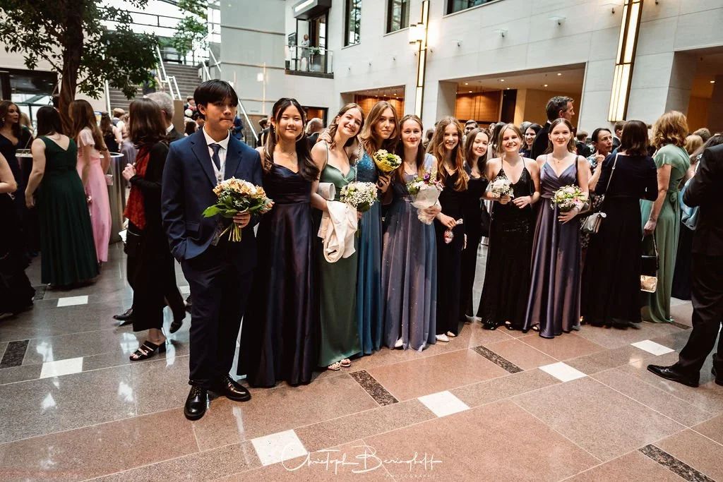 Gruppe junger Menschen in eleganten Kleidern und Anzügen beim Empfang in einem modernen, hellen Gebäude, einige halten Blumen.