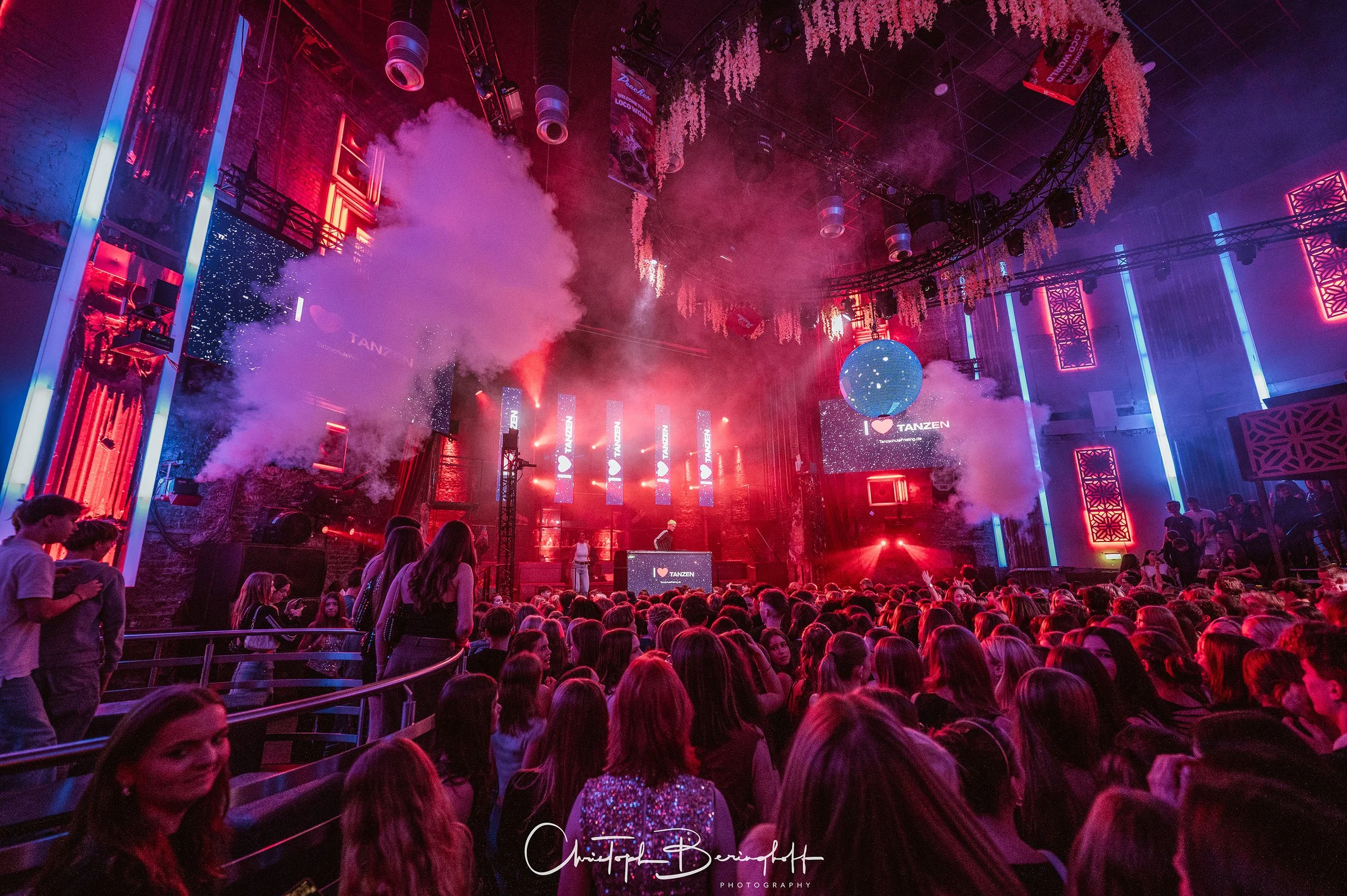 Menschenmenge tanzt bei einem Musik-Event in einem dunklen, mit Neonlichtern beleuchteten Raum mit Rauch und Discokugel an der Decke.