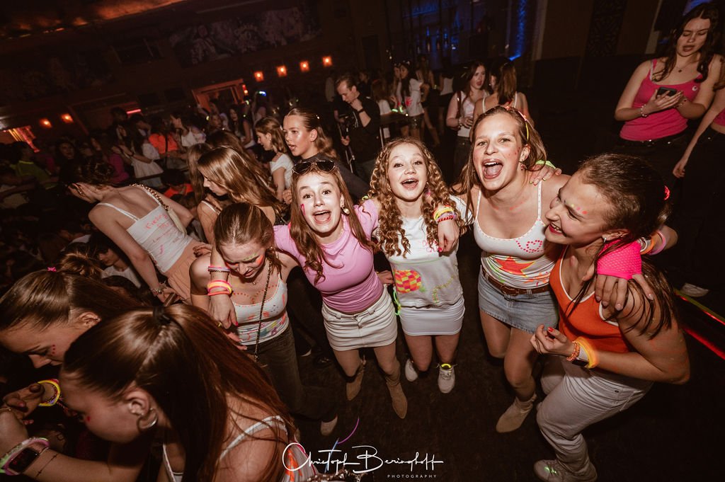 Young women are dancing and laughing at a party in a dark, festively lit setting.
