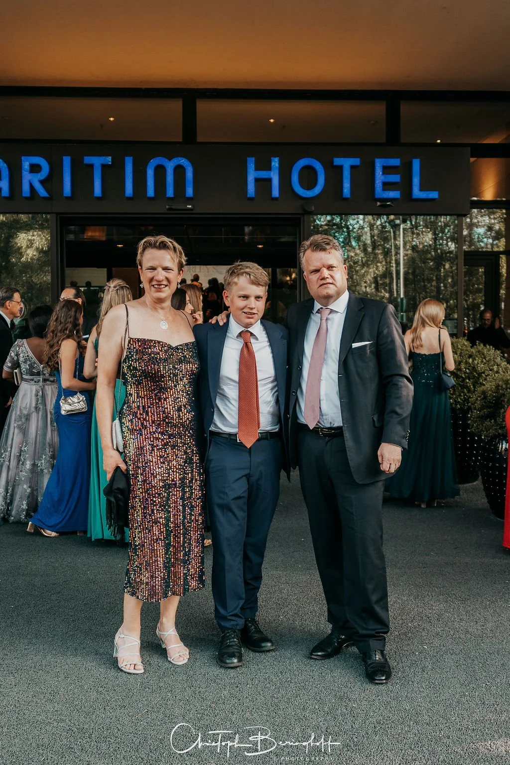 Drei Personen in formeller Kleidung stehen vor dem Eingang des Aritim Hotels bei einer Veranstaltung.