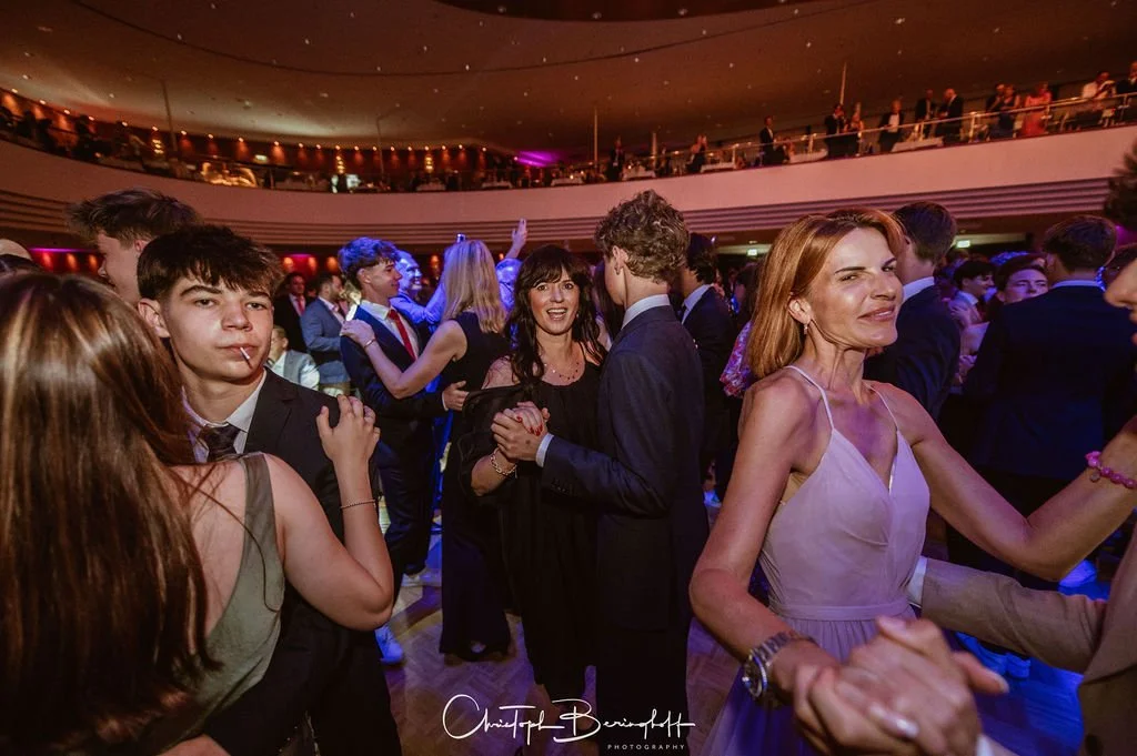 Menschen tanzen und feiern in einem festlichen Saal, viele tragen formelle Kleidung, einige lachen und haben Spaß.