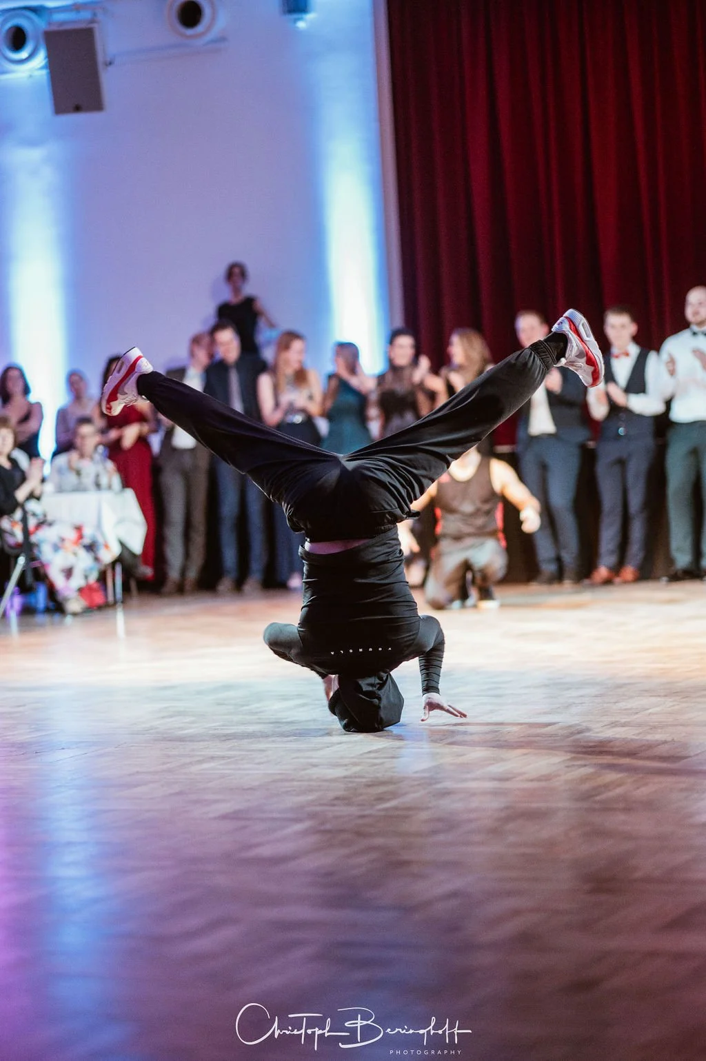 Ein Tänzer führt einen Breakdance-Move auf einer Bühne vor, mit einem Publikum im Hintergrund.