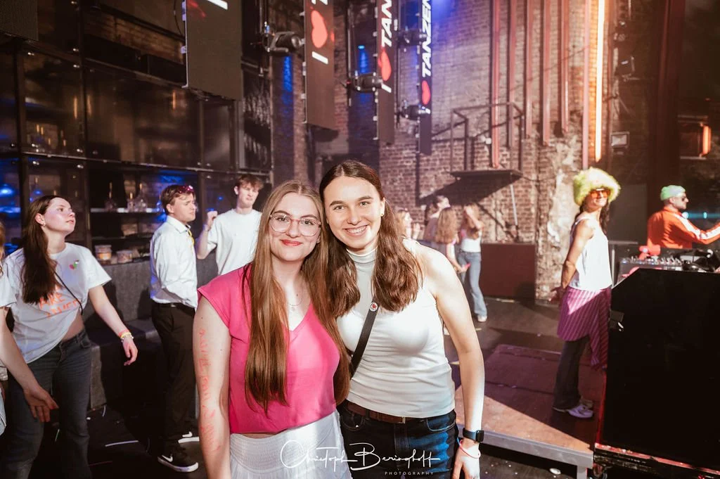 Two smiling young women are posing in a club with other people in the background who are dancing and working at the DJ booth.