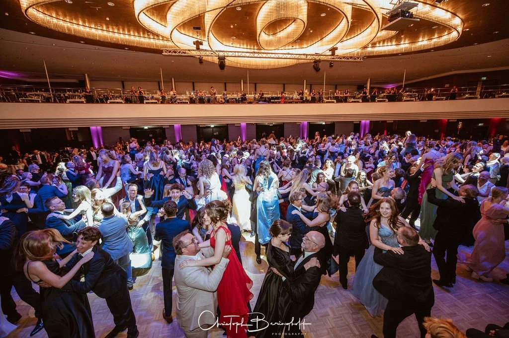 Menschen tanzen auf einer großen, gut beleuchteten Ballsaalveranstaltung, viele in eleganten Abendkleidern und Anzügen, mit einer mehrstöckigen Tanzfläche und einem modernen, dekorativen Lichtdesign an der Decke.