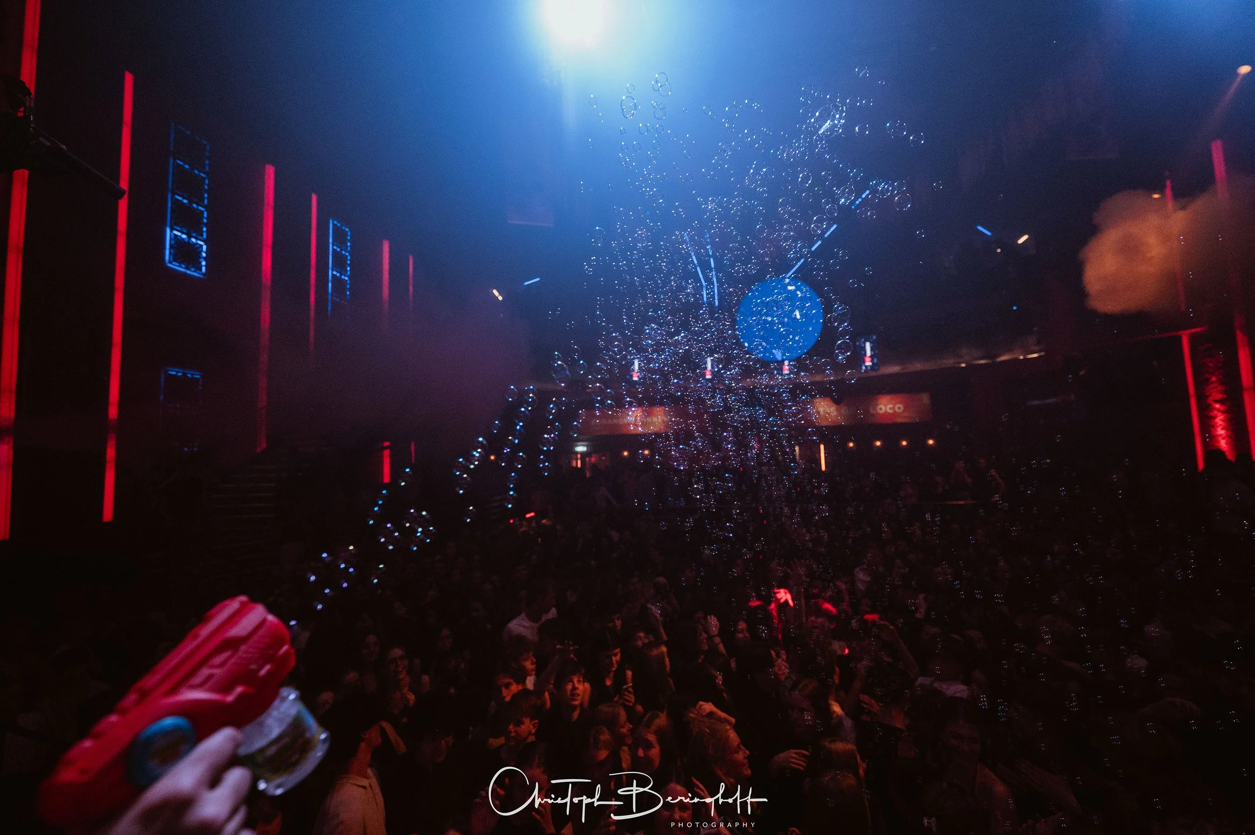 Menschenmenge in einem dunklen Club mit bunten Lichteffekten, einer Wasser- und Blaseninstallation in der Mitte, die von oben zu sehen ist. Im Vordergrund hält jemand einen roten Wasserpistolen. Die Atmosphäre ist lebhaft und festlich.