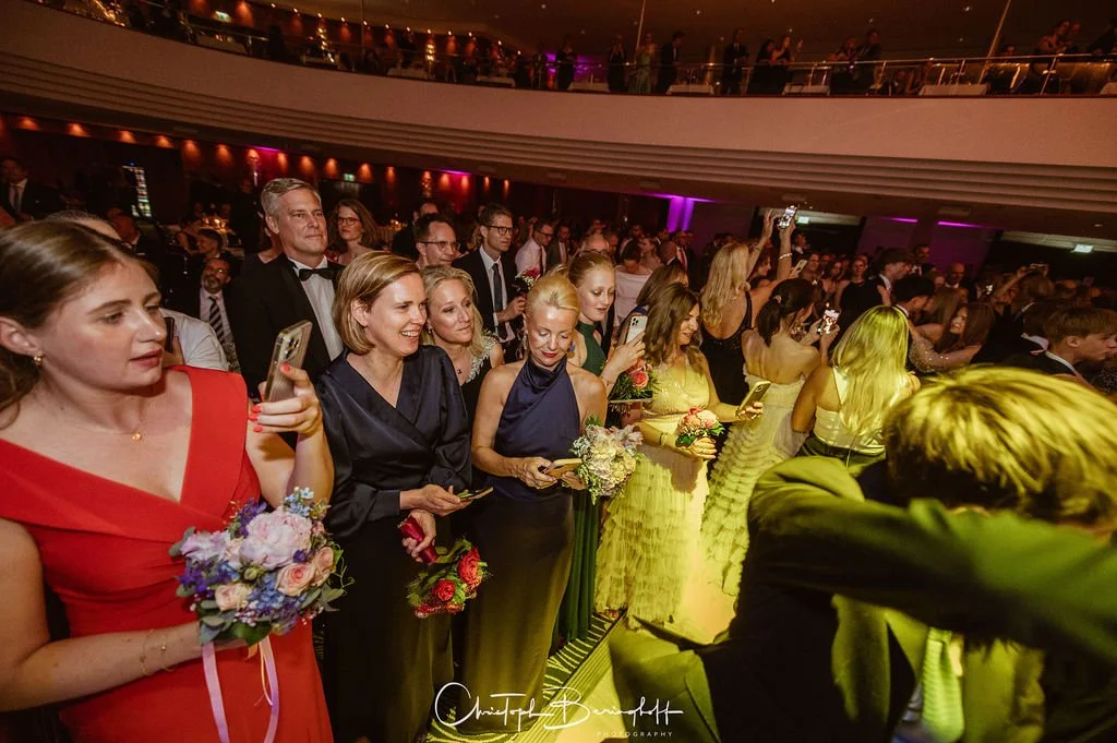 Menschen bei einer formellen Veranstaltung, einige halten Blumen und nehmen Fotos auf, im Hintergrund eine mehrstöckige Veranstaltungsstätte mit Kerzen und bunten Lichtern.