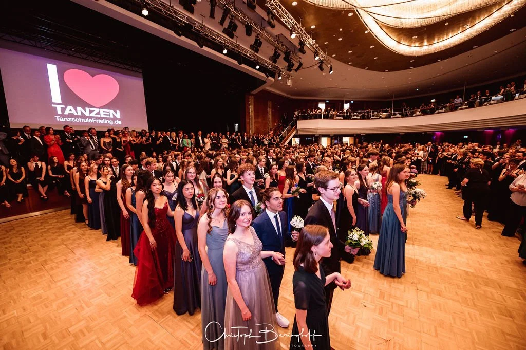 Junge Menschen in formeller Kleidung beim Abschlussball in einem großen Saal mit Bühne und einer großen Projektion, die 'I ♥ TANZEN' zeigt.