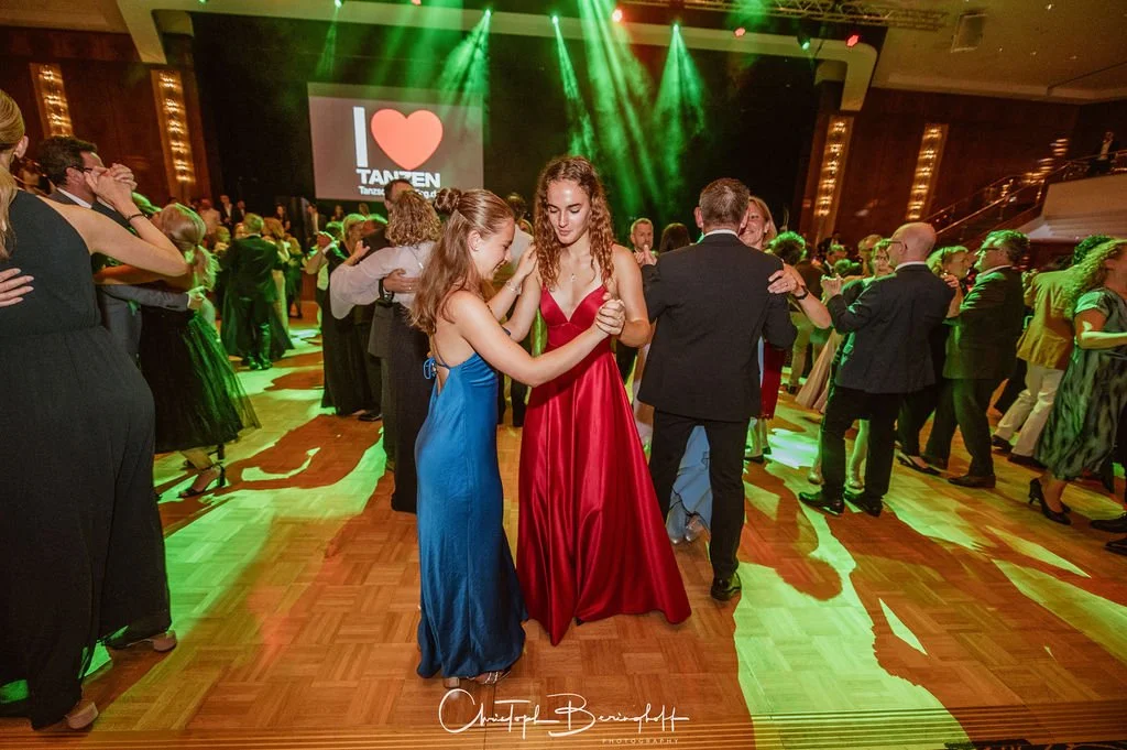 Paare tanzen auf einer festlichen Tanzveranstaltung mit bunten Lichtern und einer großen Bühne im Hintergrund.