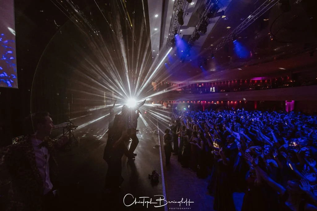 Tanzparty in einem dunklen, gut beleuchteten Saal, eine Person tanzt vor einem spektakulären Lichtblitz, die Menge applaudiert und hat die Hände erhoben.