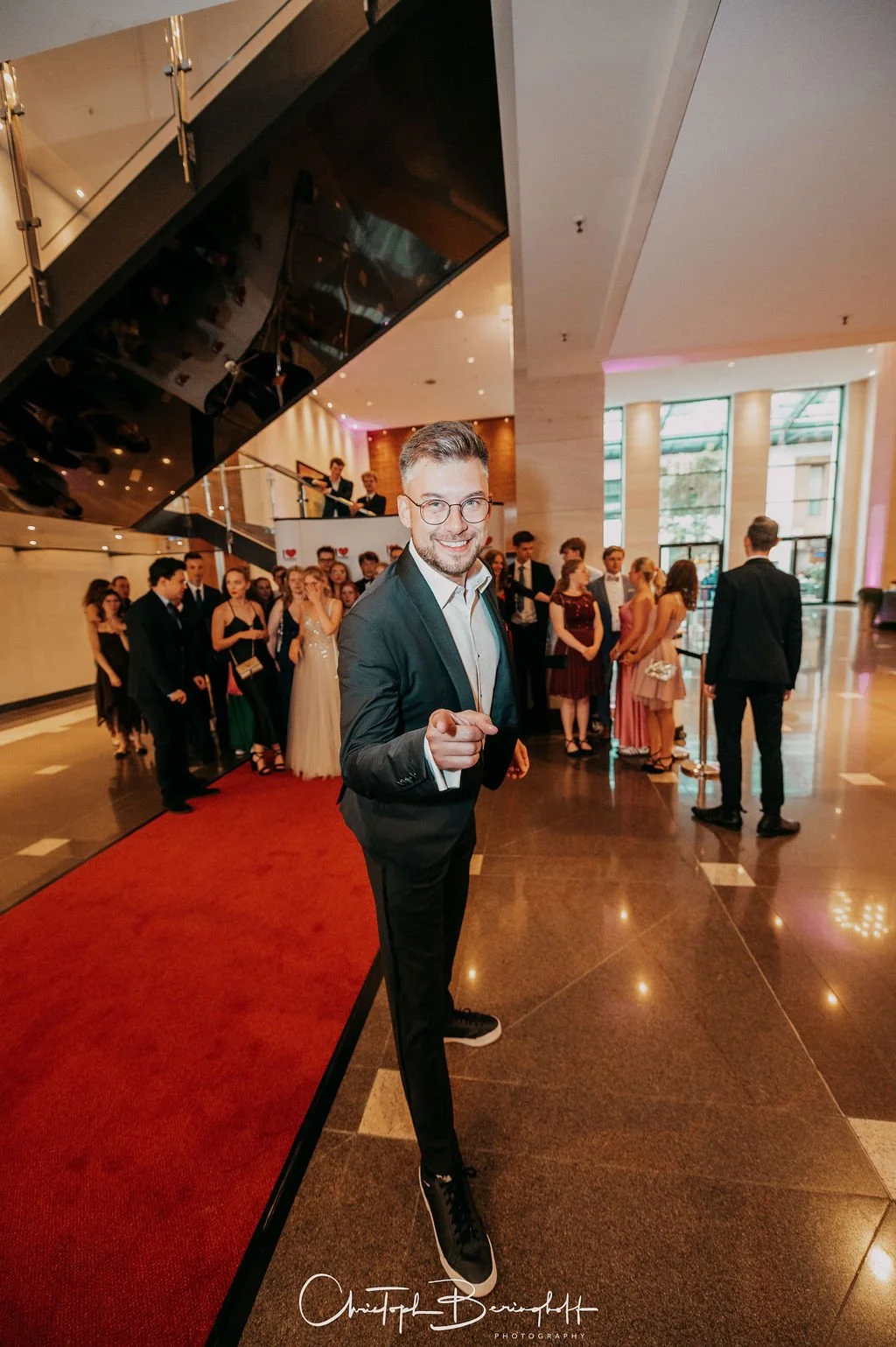 Ein glücklicher Mann in formeller Kleidung, der auf einer roten Teppich im Eingangsbereich eines festlichen Events steht, umgeben von einer Menschenmenge in eleganter Kleidung in einem modernen Hotel-Lobby mit großen Fenstern und eleganten Dekoration