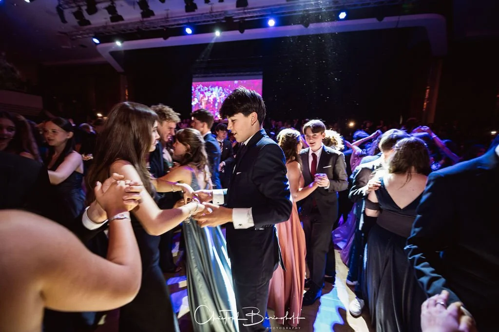 Jugendliche tanzen auf einem festlichen Ball in formeller Kleidung, umgeben von dunkler Ambiente und buntem Licht.