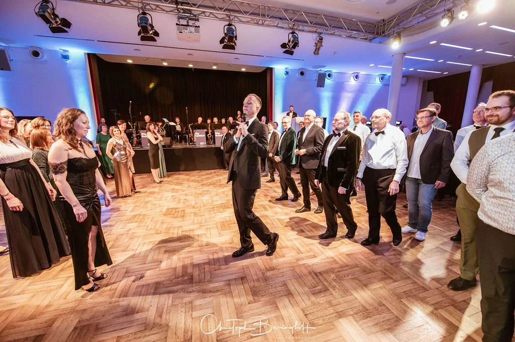 Menschen tanzen und feiern in einem festlich dekorierten Raum mit einer Live-Orchester-Band auf der Bühne.