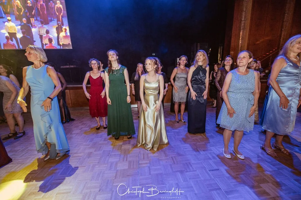 Mehrere Frauen tanzen auf einer Tanzfläche in eleganten Kleidern bei einer Feier oder einem Ereignis.