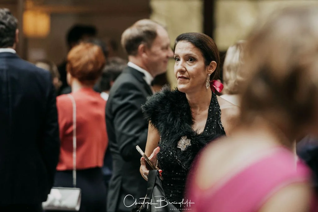 Frau in schwarzem Abendkleid und roten Haaren bei einem formellen Event, umgeben von anderen Gästen in eleganter Kleidung.