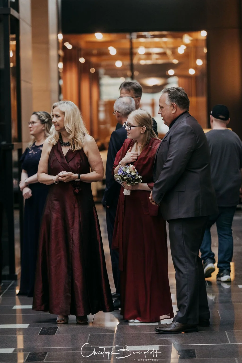 Gruppe von Menschen in formeller Kleidung, eine Frau hält einen Blumenstrauß, sie befinden sich in einem eleganten Innenraum bei einer Veranstaltung.