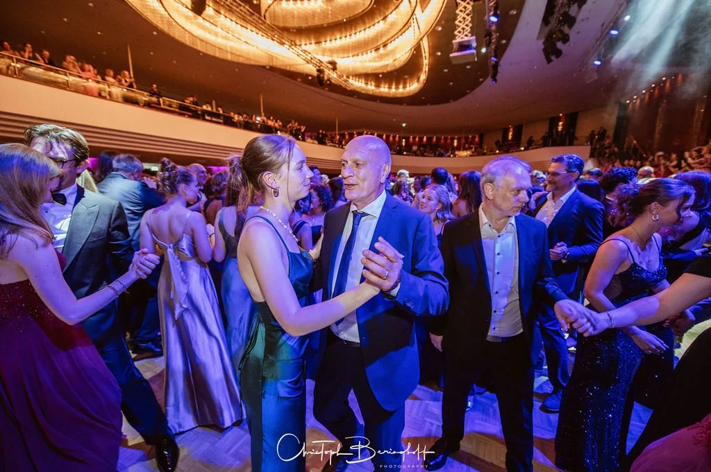 Paare tanzen in festlicher Abendgarderobe in einem eleganten Saal mit goldener Lichtdecke bei einer formellen Veranstaltung.