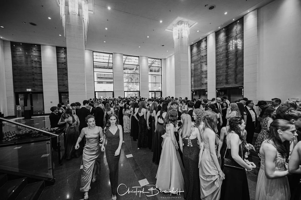Menschen in eleganten Abendkleidern und Anzügen bei einer formellen Veranstaltung in einem großen, modern eingerichteten Saal mit hohen Fenstern und Dekorationen.