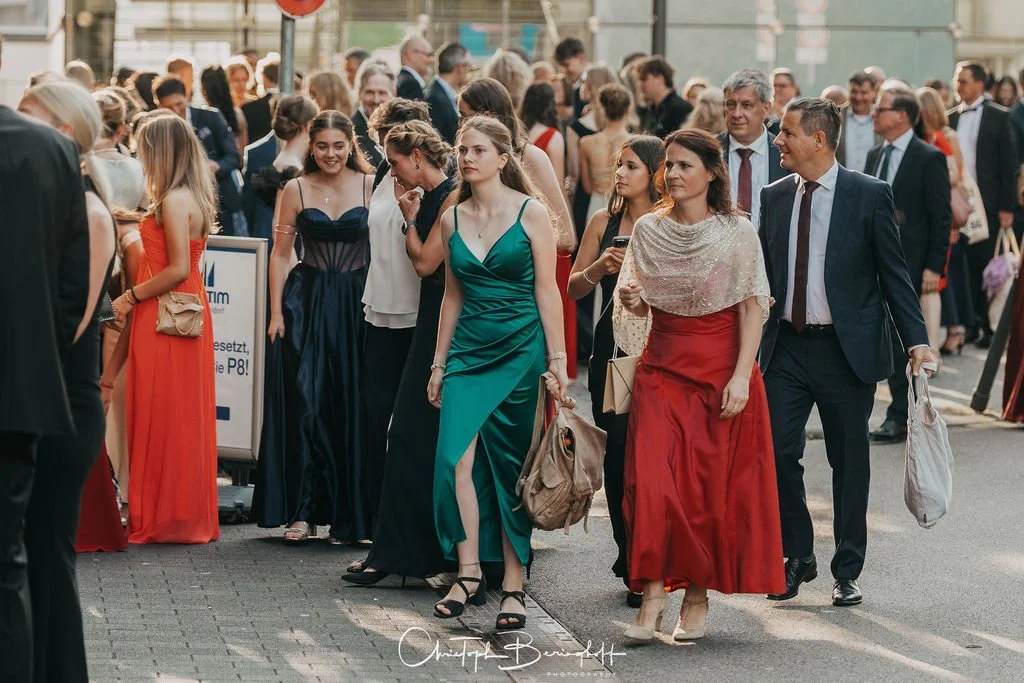 Menschen in formeller Kleidung auf einer Straßenveranstaltung, viele Frauen in eleganten Abendkleidern, Männer in Anzügen.
