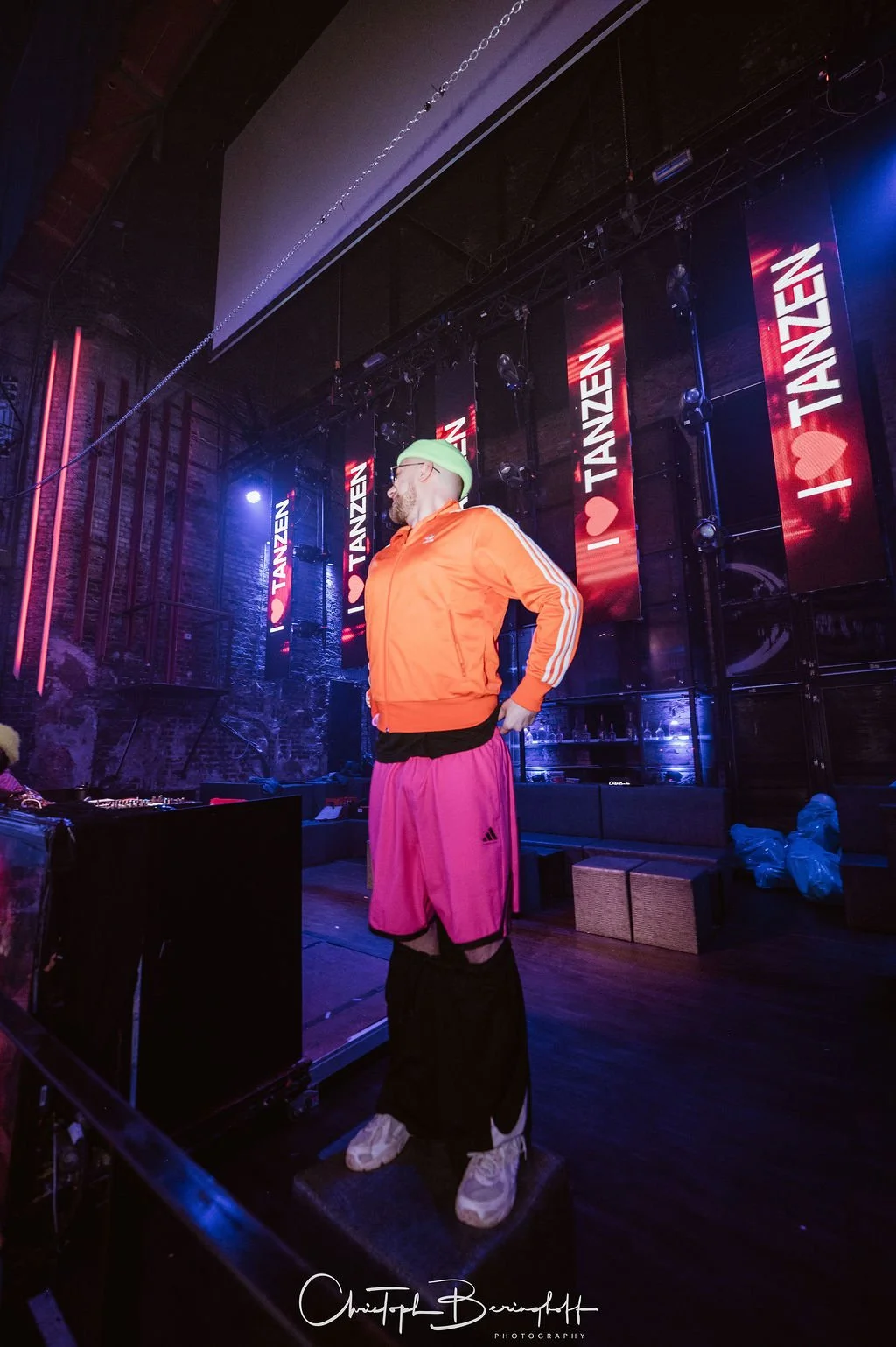 A man is standing on a stage lit by neon lights, wearing an orange jacket, pink shorts, black pants, and white sneakers; in the background are banners that read "I ❤️ DANCING."