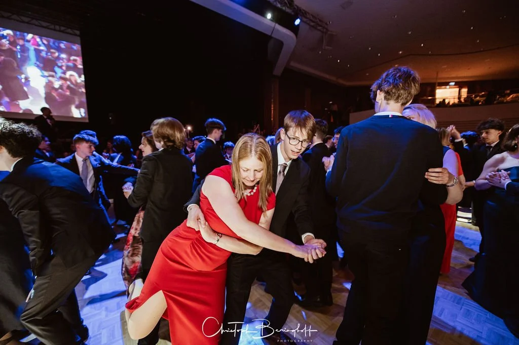 Menschen tanzen auf einer Party oder einem Ball, in der Mitte eine lachende Frau in rotem Kleid, begleitet von einem Mann in Anzug.