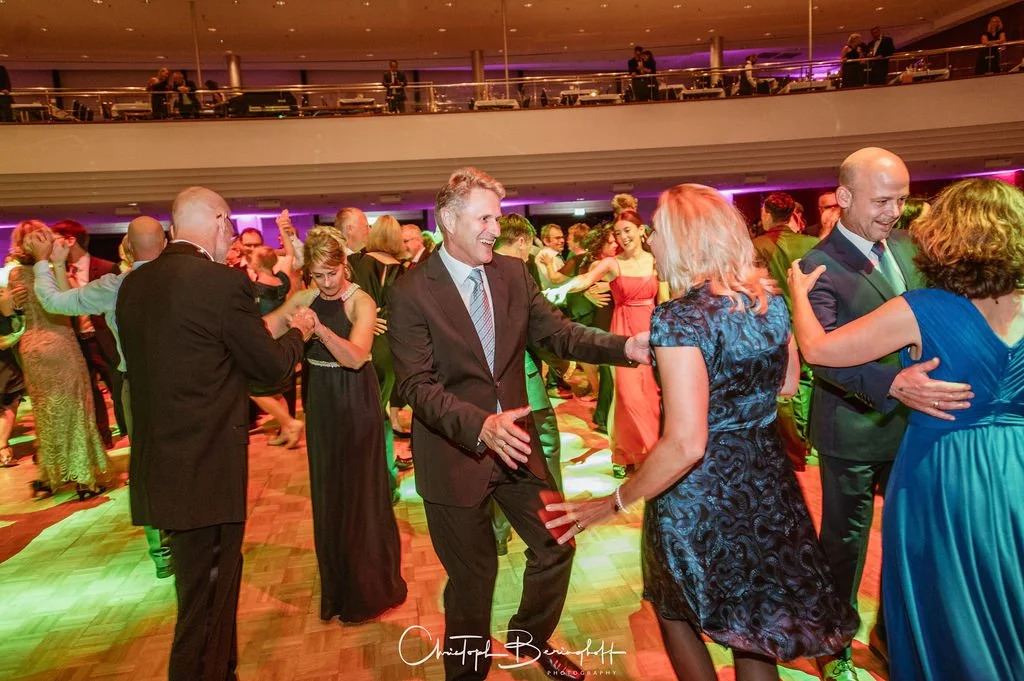 Paare tanzen auf einer Gala im Ballsaal, viele tragen elegante Abendkleidung, während die Gäste im Hintergrund auf einer oberen Etage zuschauen.