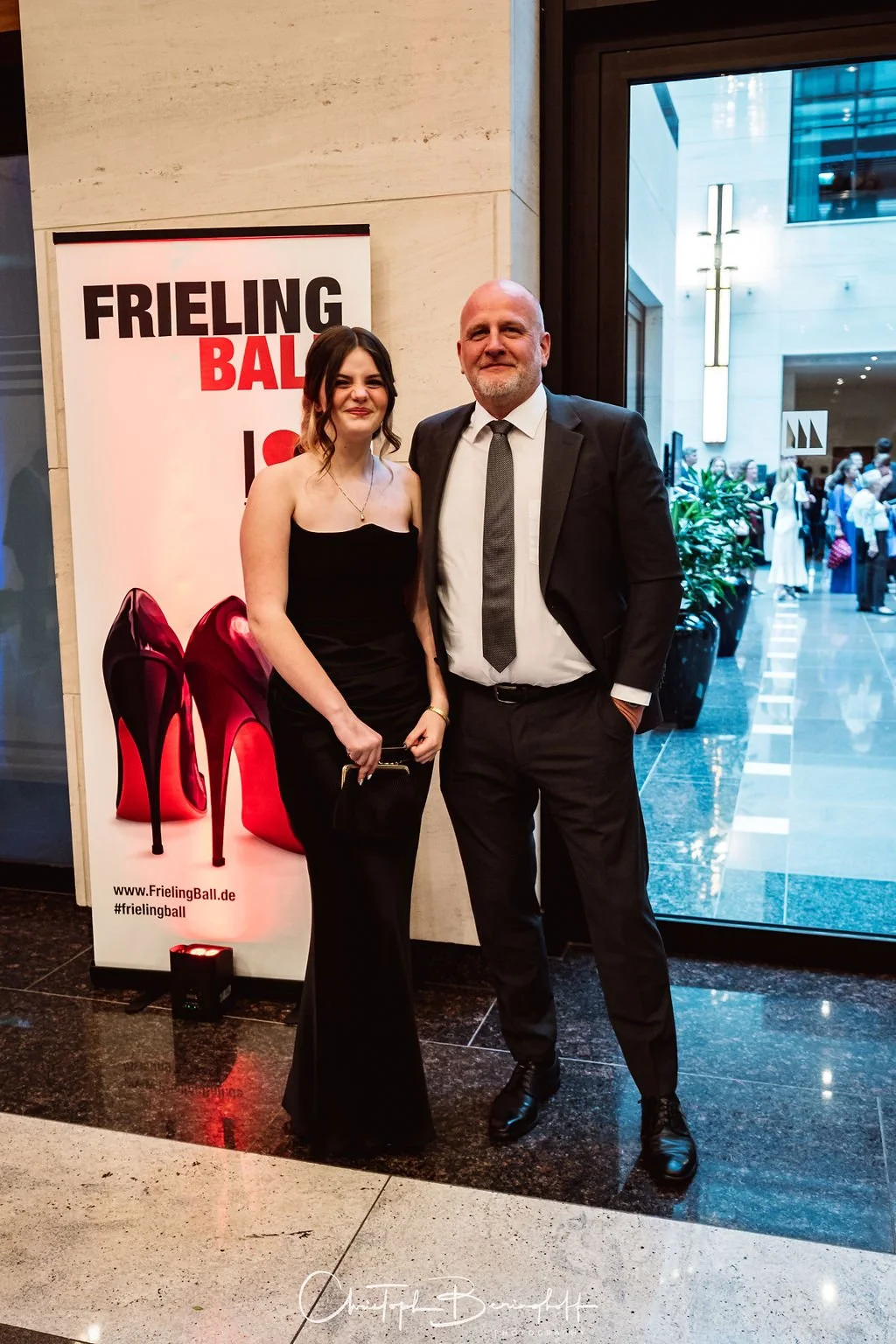 Ein Mann und eine Frau stehen nebeneinander vor einem Poster mit roten High Heels bei einem Ball. Im Hintergrund sind Menschen in einem modernen Gebäude sichtbar.