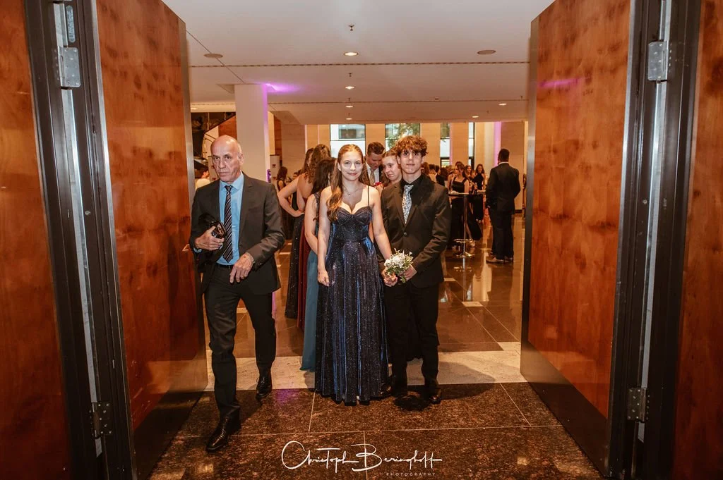 Junge Frau und Mann im eleganten Kleid und Anzug, Hand in Hand, bei einer formellen Veranstaltung, umgeben von weiteren Gästen im Hintergrund.