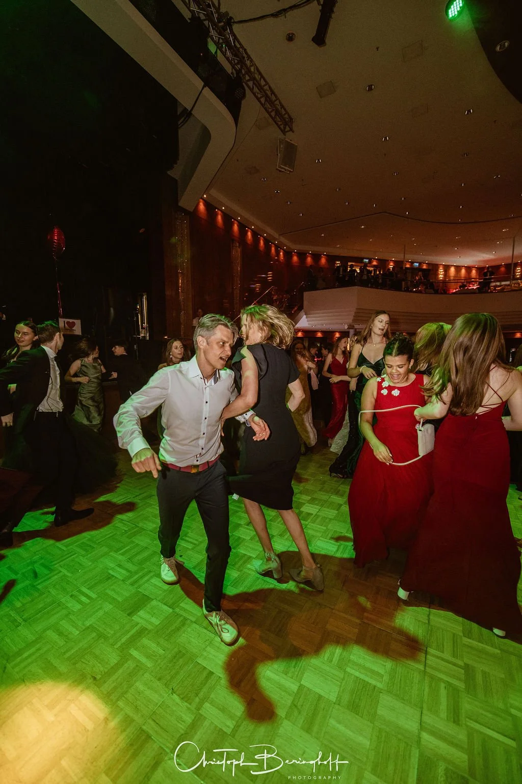 Menschen tanzen auf einer Party, viele tragen elegante Kleidung, in einem festlich dekorierten Raum mit Tanzfläche und Bühne.