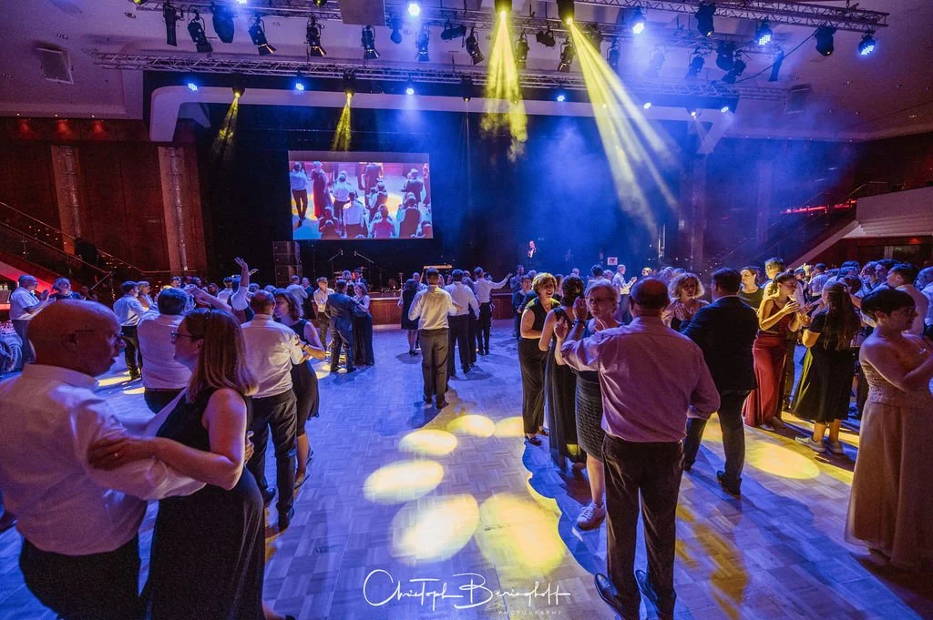 Tanzende Menschen bei einer Tanzveranstaltung in einem großen Saal mit Bühnen- und Lichttechnik.