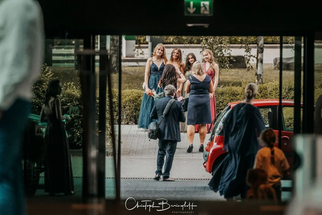 Aufnahme von einer Gruppe junger Frauen, die draußen posieren, während andere Personen im Vordergrund und daneben unterwegs sind.