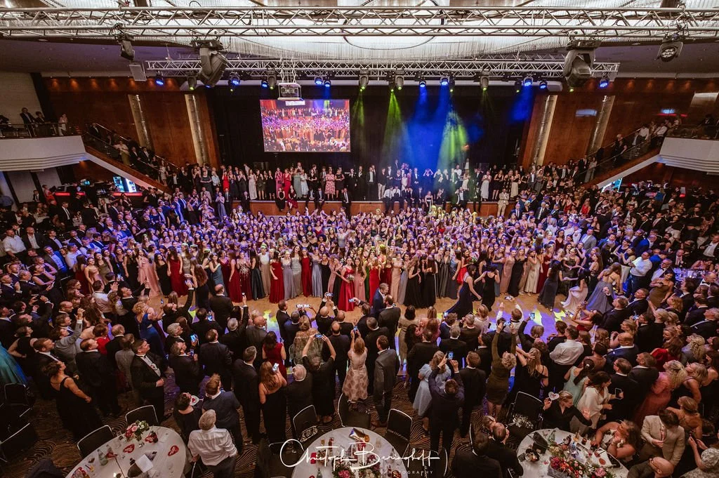 Große Ballsaal mit vielen Feiernden, Tanzfläche in der Mitte, Bühne im Hintergrund, bunte Lichter, Menschen in formeller Kleidung, festlicher Anlass.