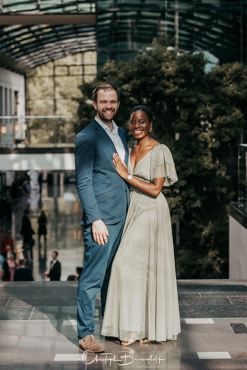 Ein Mann in einem blauen Anzug und eine Frau in einem langen, hellgrünen Kleid stehen nebeneinander und lächeln vor einem modernen Gebäude.