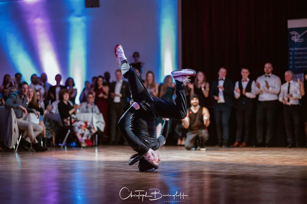 Ein Tanzmeister führt einen Breakdance-Move auf einem Parkettboden vor, während ein Publikum aus Gästen im Hintergrund zuschaut und applaudiert.
