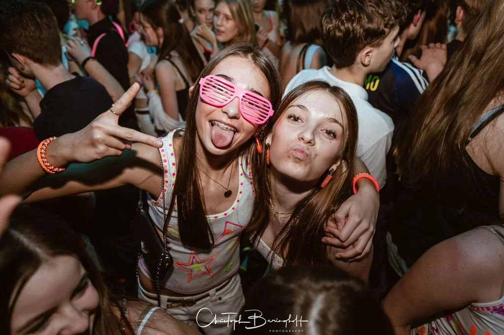 Two young women are partying; one is wearing pink sunglasses, both are happy and hugging each other, with many other partygoers in the background.