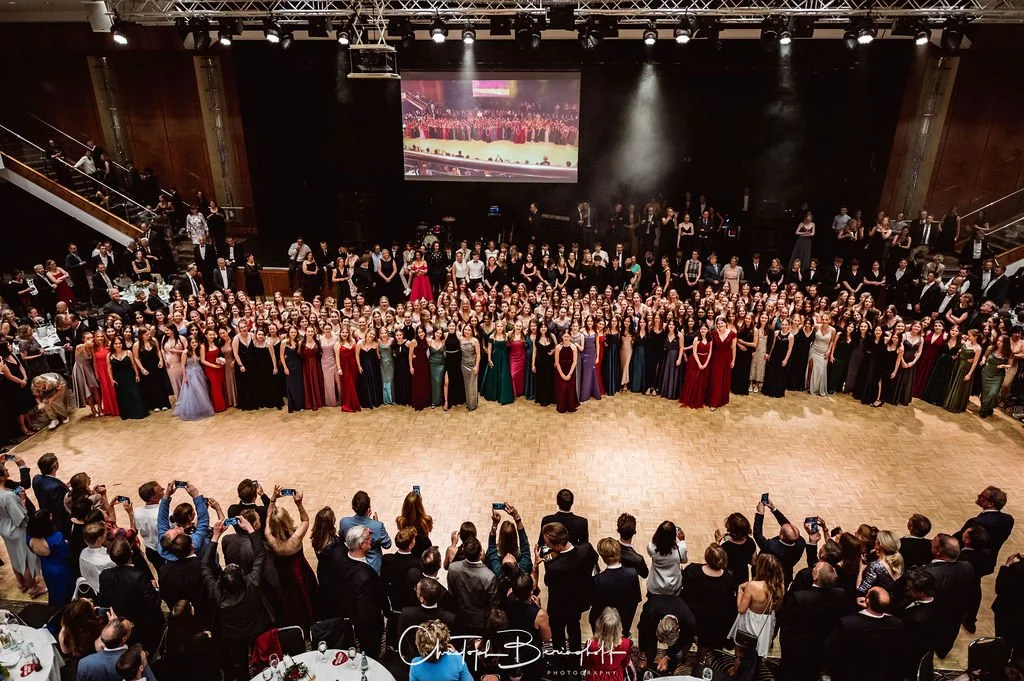 Eine große Gruppe von Tänzern in formellen Abendkleidern und Anzügen posiert für ein Foto in einem Festsaal, während Gäste sie umzingeln und Fotos machen.