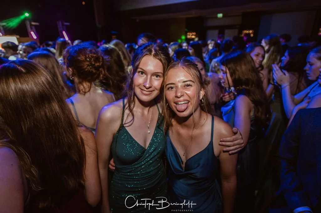 Zwei junge Frauen tanzen und lachen auf einer Party mit vielen Gästen, bunte Lichter im Hintergrund.