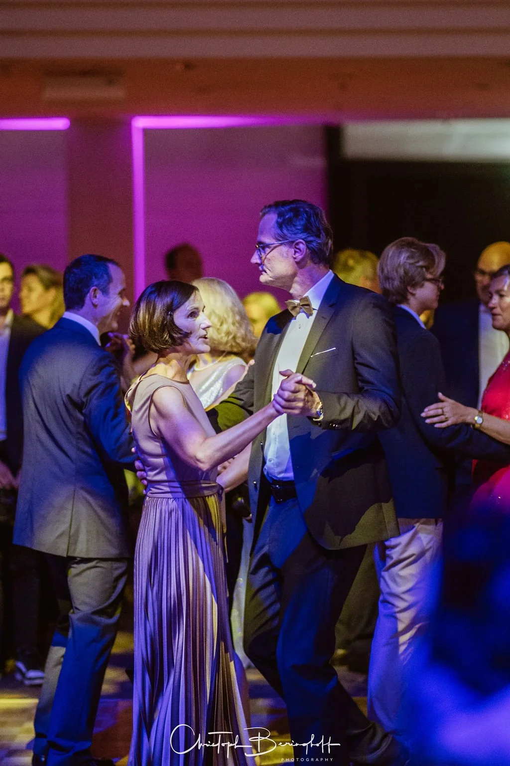 Paare tanzen auf einer eleganten Party im Abendlicht, Männer in Anzügen und Frauen in schicken Kleidern.