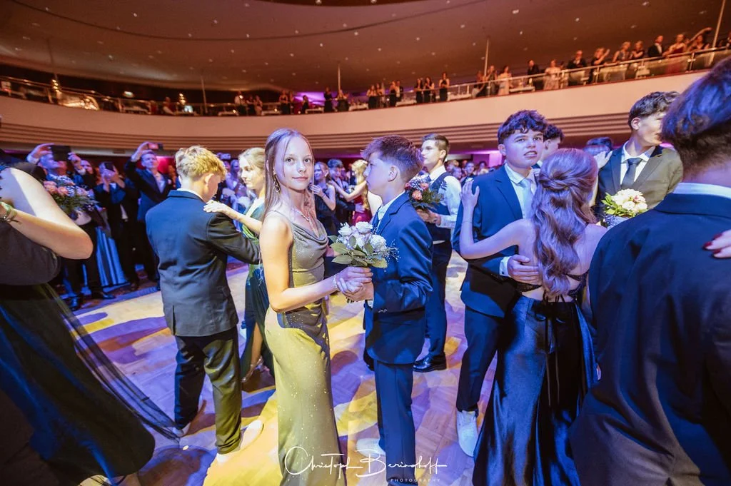 Jugendliche tanzen bei einem festlichen Ball im eleganten Abendoutfit auf einem Tanzparkett in einem großen Saal mit Publikum im Hintergrund.