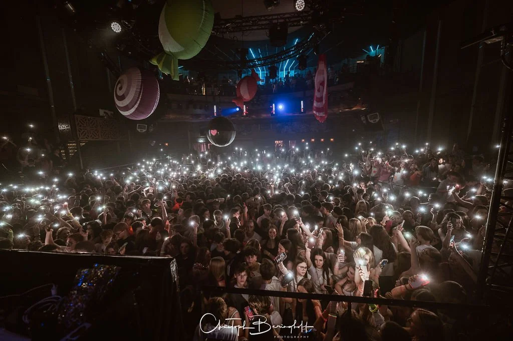 People in a dark club setting taking photos with their cell phones; lots of lights are shining.