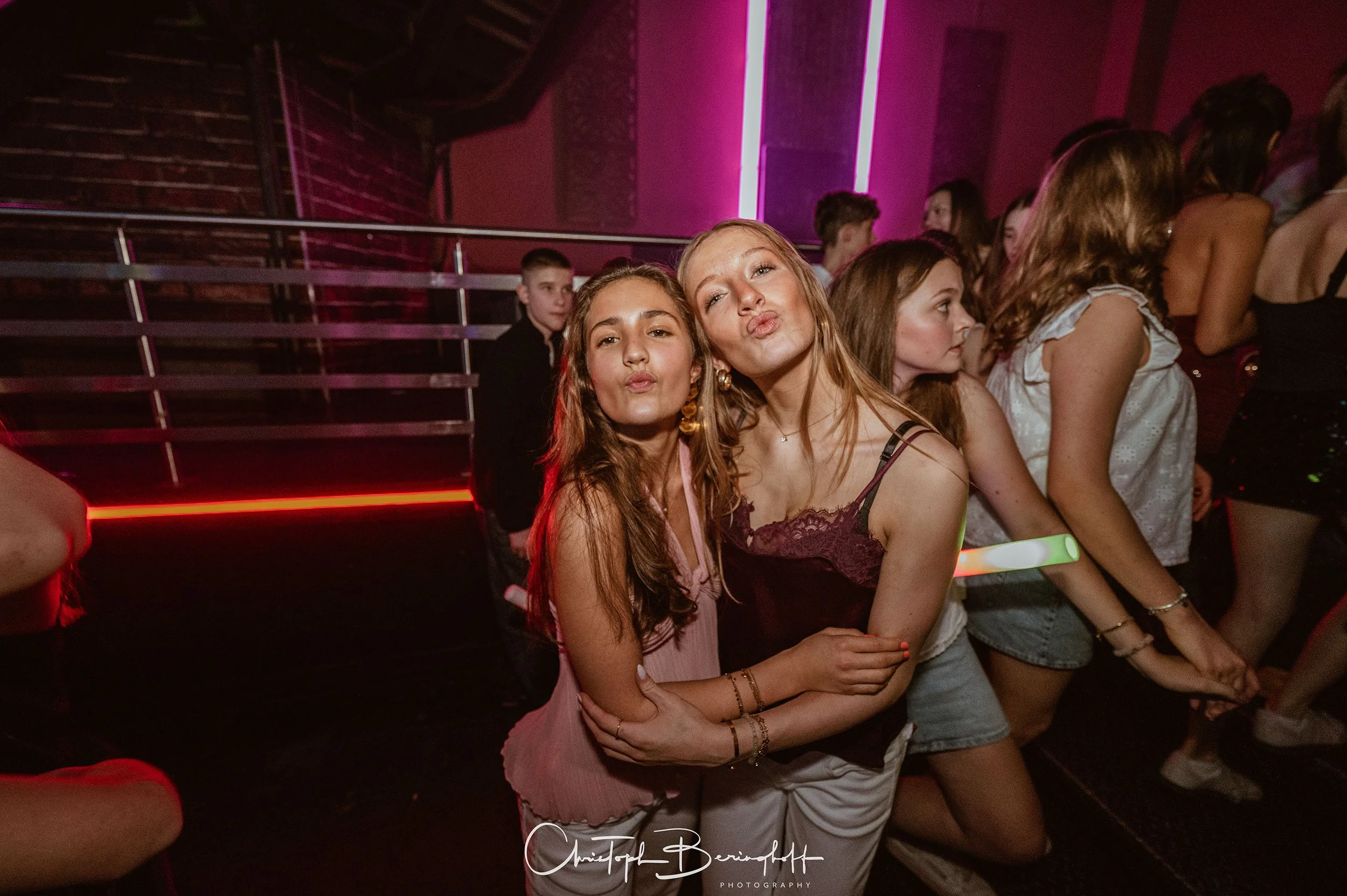 Zwei lachende Mädchen posieren bei einer Party in einem dunklen Raum mit Rot- und Pink-Lichtern.