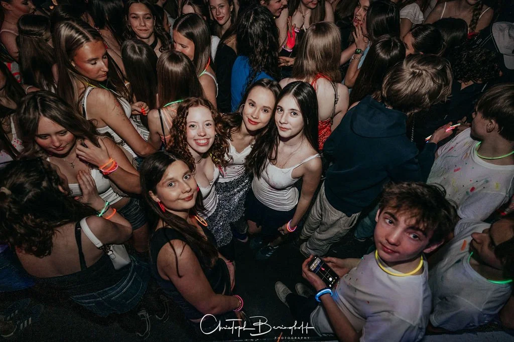 A group of young people are dancing at a party; many are wearing neon wristbands. In the center are three girls looking at the camera.