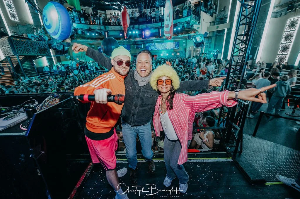 Three cheerful people are celebrating near a DJ booth in a large, dark venue, with colorful lights and an audience in the background.