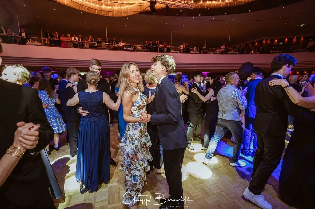 Menschen tanzen auf einer Party im festlichen Rahmen, im Vordergrund ein Paar, das sich küsst und tanzt.